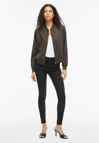 Bomberjakke i mørkebrunt stoff med glatt finish, glidelåsfeste foran og to sidelommer; kombinert med svarte skinny jeans og svarte høye hæler.