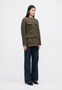 Brązowa marynarka w kratę z czterema przednimi kieszeniami, okrągłym dekoltem i czarnymi guzikami, w połączeniu z ciemnymi spodniami z szerokimi nogawkami i czarnymi butami.