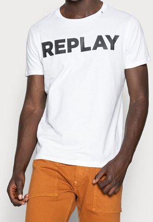 Personne à la peau foncée portant un t-shirt blanc avec « REPLAY » en lettres noires et un pantalon orange brûlé avec des poches avant.