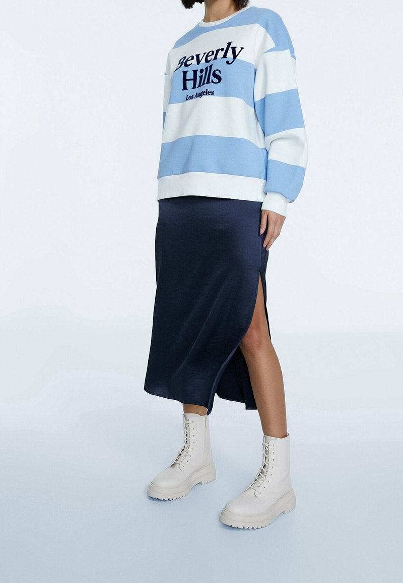 Personne portant un sweat-shirt rayé bleu et blanc avec le texte « Beverly Hills », une jupe fendue bleu marine et des bottes à lacets écru.
