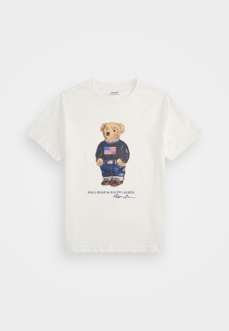 T-shirt branca de manga curta com um gráfico de um urso a usar um suéter azul marinho com a bandeira americana, jeans de ganga e sapatos castanhos.