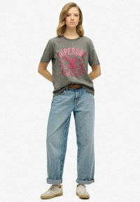 Camiseta de algodón gris con estampado gráfico en rosa, combinada con unos pantalones vaqueros de cintura alta de tiro corto y con dobladillo enrollado en azul claro. La modelo lleva zapatillas beige.