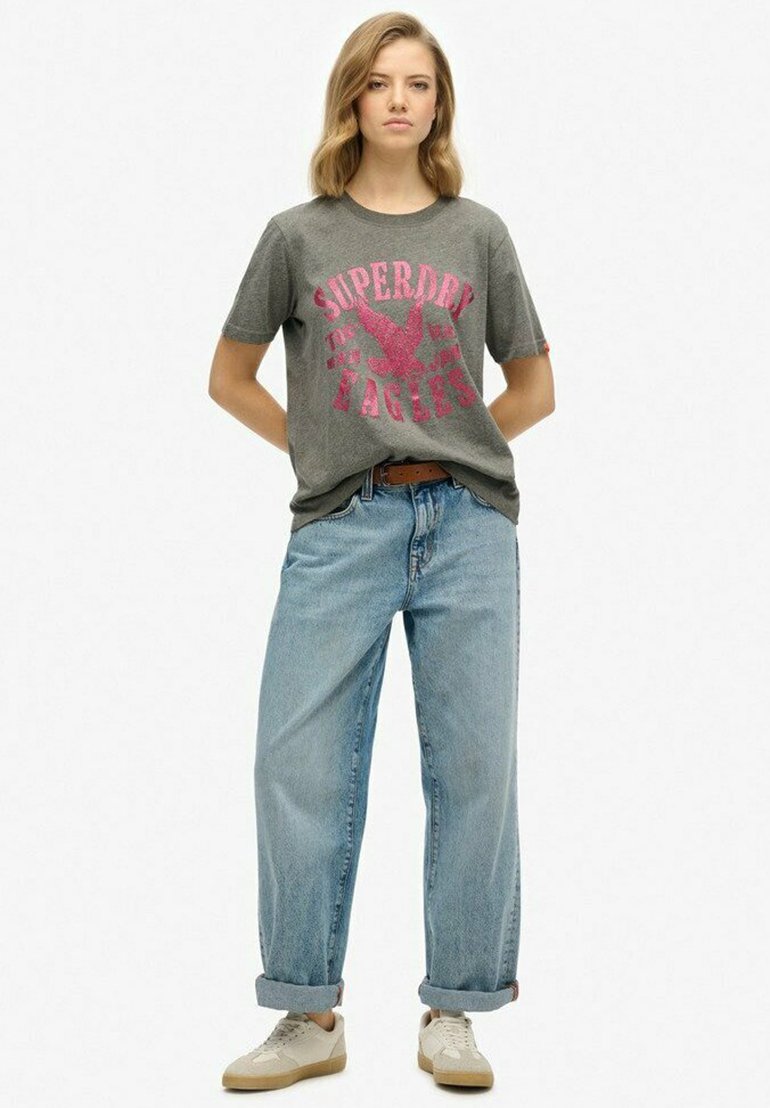 Camiseta de algodón gris con estampado gráfico en rosa, combinada con unos pantalones vaqueros de cintura alta de tiro corto y con dobladillo enrollado en azul claro. La modelo lleva zapatillas beige.
