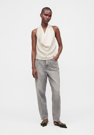 Femme portant un haut crème sans manches drapé, un jean gris ample et des ballerines noires pointues, debout avec une main dans la poche sur fond blanc.