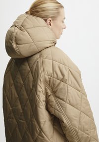 Beige vuorattu puffer-takki, jossa on suuri pyöreä huppu ja timanttikuosi, sileä ja läpinäkymätön kangas ja näkyvät tikkauskorostukset.