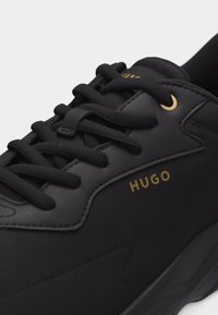 Μαύρο αθλητικό παπούτσι με μαύρα κορδόνια, λεπτομέρεια χρυσής οπής και χρυσό λογότυπο "HUGO" στο πλάι, που δείχνει συνδυασμό υφής και λείας υφάσματος.