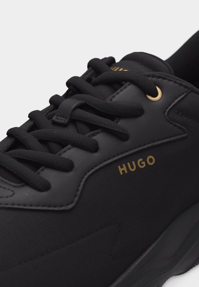 Baskets noires avec lacets noirs, détails œillets dorés et logo "HUGO" doré sur le côté, affichant une combinaison de tissu texturé et lisse.