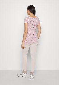 Light pink short-sleeve top with black floral patterns, paired with beige leggings. The model wears silver sneakers and light gray ankle socks.