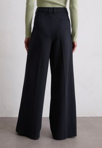 Schwarze weite Hosen aus einem strukturierten Stoff, mit hoher Taille und dezenten Falten, sowie einem Detail mit einer Gesäßtasche.