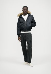 Ναυτική βομβιστική ζακέτα με επένδυση από γούνινη κουκούλα, κλείσιμο με φερμουάρ, φαρδιά cargo παντελόνια, φορεμένο με λευκό μπλουζάκι και ελαφριά μπεζ αθλητικά παπούτσια.