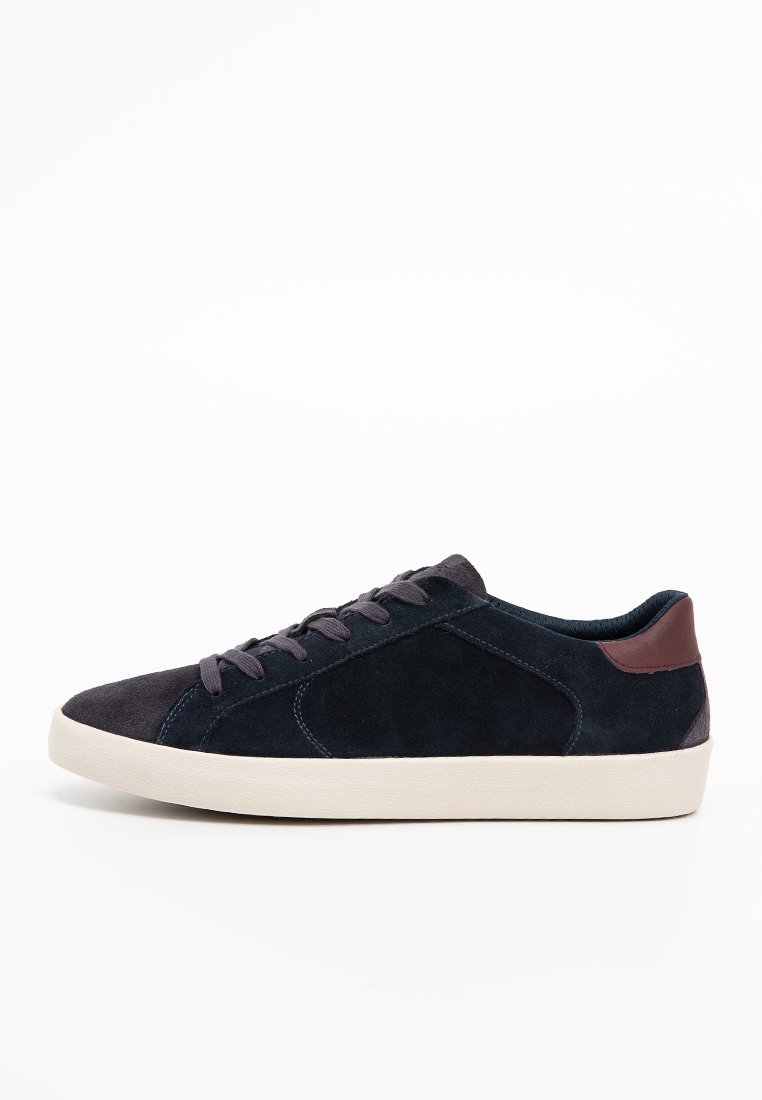 Sneaker en suede bleu marine avec une semelle en caoutchouc crème, bout rond et accent de talon marron foncé. Présente des lacets plats et des détails de couture subtils.