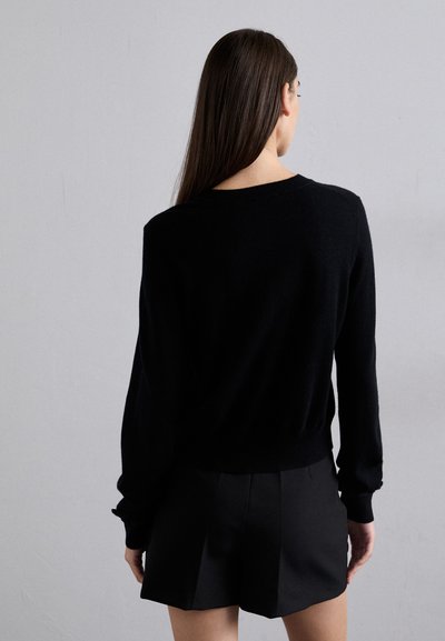 Maglione nero lavorato a maglia con scollo rotondo e maniche lunghe. Il design presenta polsini a coste e un orlo aderente. Indossato con pantaloni corti neri.