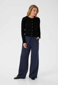 Cardigan noir avec fermeture à boutons, associé à un pantalon ample en bleu foncé, texturé. Le modèle se tient avec une main dans la poche.