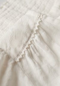 Strukturiertes, weißes Gewebe mit zarter Stickerei und einem Rüschensaum, das ein weiches, leichtes Material mit dezenten Mustern präsentiert.