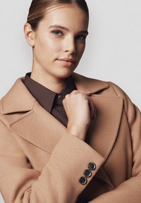 Jasnobrązowy płaszcz wełniany z szerokimi zakonami, z trzema czarnymi guzikami na mankietach, noszony na ciemnobrązowej koszuli. Gładka tekstura i dopasowany krój.