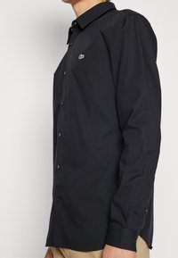 Camisa negra abotonada con mangas largas, tela texturizada y un pequeño logo verde en el pecho. Corte ligeramente entallado.