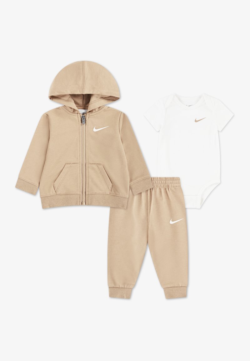Sweat à capuche beige zippé avec poches avant, pantalon de survêtement beige assorti et body à manches courtes blanc, tous arborant le logo Nike.
