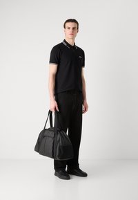 Čierna polokošeľa s bielym prúžkovaným golierom, čierne nohavice, drží čiernu športovú tašku s embosovaným logom. Hladká tkanina a minimalistický dizajn.