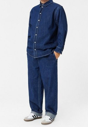 Dunkelblaue Denim-Bluse mit Knopfleiste, kombiniert mit weit geschnittenen dunkelblauen Denim-Hosen. Getragen mit grauen und weißen Sneakern, die Streifen haben.