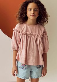 Helles rosa Oberteil mit puffigen Ärmeln und Rüschensaum, kombiniert mit blauen gestreiften Short-Hosen mit floralem Stickmuster auf den Taschen.