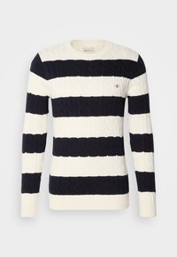 Pull ras-du-cou à larges rayures horizontales en maille torsadée noire et blanche, avec un petit logo sur la poitrine, côté gauche.