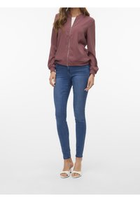 Donkerrode bomberjack met een ritssluiting aan de voorkant, een aansluitend ontwerp en geribbelde manchetten, gecombineerd met blauwe skinny jeans en witte open-toe hakken.