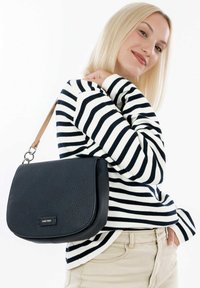 Black pebbled leather shoulder bag with a curved flap and tan leather strap. Striped long-sleeve top and light-colored pants are worn.