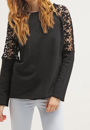 Vrouw draagt een zwarte top met lange mouwen met bloemenkant aan de mouwen en lichtgrijze broek, staand tegen een effen achtergrond.