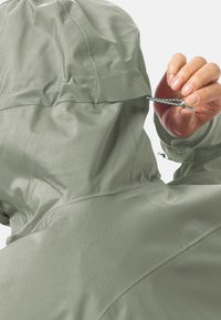 Lichtgroene waterdichte jas met een capuchon, gemaakt van een gestructureerde stof en met een aan te passen koord aan de achterkant.