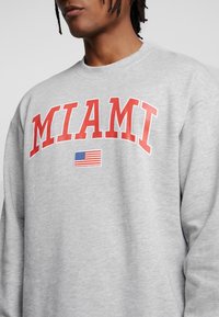 Sweatshirt gris avec "MIAMI" imprimé en lettres rouges en arc ; détail de petit drapeau américain en dessous du texte ; col rond et coupe décontractée.