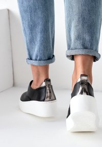 Black leather slip-on shoes with metallic accent heel and thick white sole; denim jeans rolled at the ankles, showcasing the shoes.
