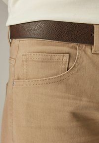 Close-up van beige broeken met gedetailleerde stiknaden en een gestructureerde donkerbruine leren riem om de taille.
