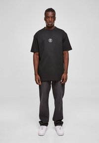 Juodas marškinėlis su apvaliu baltu logotipu ant krūtinės, derinamas su tamsiai mėlynomis džinsinėmis kelnėmis ir baltais sportbačiais, pasižymintis laisvu siluetu ir trumpomis rankovėmis.