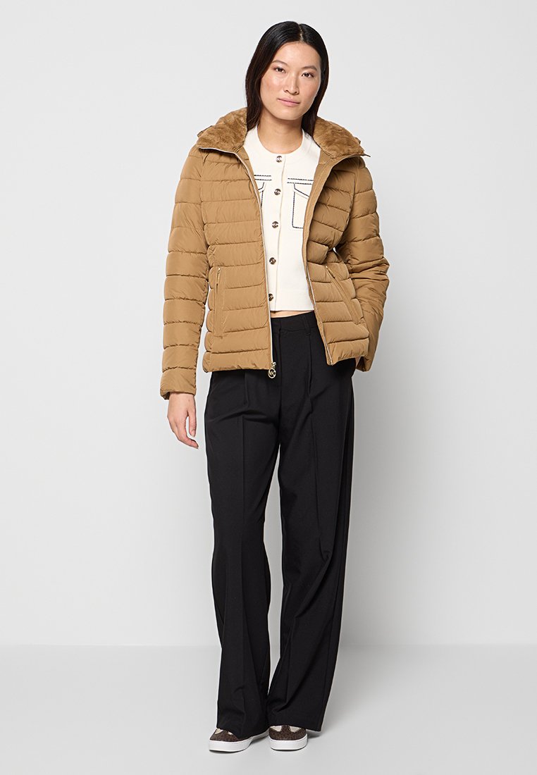 Braune Daunenjacke mit Kapuze aus Kunstfell, gestepptem Design, Frontreißverschluss, kombiniert mit einem weißen Shirt und schwarzen Hosen, die eine lockere Passform haben.