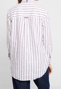 Chemise blanche avec des rayures verticales rouges, manches longues et un détail de pli au dos. Présente des fentes sur les côtés et un design de poignet à boutons.