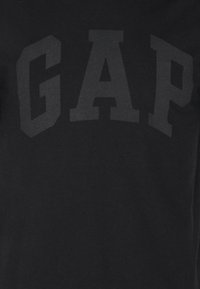 Schwarzes T-Shirt mit großem, erhabenem "GAP"-Schriftzug in einer markanten, stilisierten Schriftart. Der Stoff ist glatt mit einheitlicher Textur.