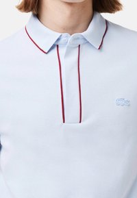 Polo-shirt bleu clair avec un piping rouge sur le col et la patte de boutonnage, porté par une personne ayant les cheveux bruns courts, logo de crocodile brodé sur la poitrine.