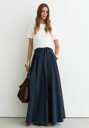 Vrouw met lang haar draagt een wit topje met korte mouwen, een marineblauwe geplooide maxi-rok met hoge taille, gouden armbanden, houdt een bruine leren tas vast, staat tegen een witte muur.