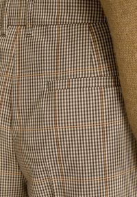Close-up van een bruine en beige geruite broekzak met subtiele oranje lijnen, gecombineerd met een textuurrijke camel-kleurige sweatermouw.