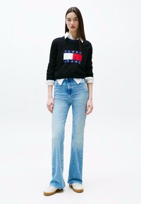 Felpa nera con logo Tommy Jeans ricamato in rosso, bianco e blu, abbinata a jeans svasati azzurri e sneakers bianche.