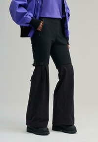 Schwarze Skihose mit ausgestellten Beinen, verstellbaren Trägern an der Taille, Reißverschlusstaschen und glatter Oberfläche, kombiniert mit schwarzen robusten Stiefeln.