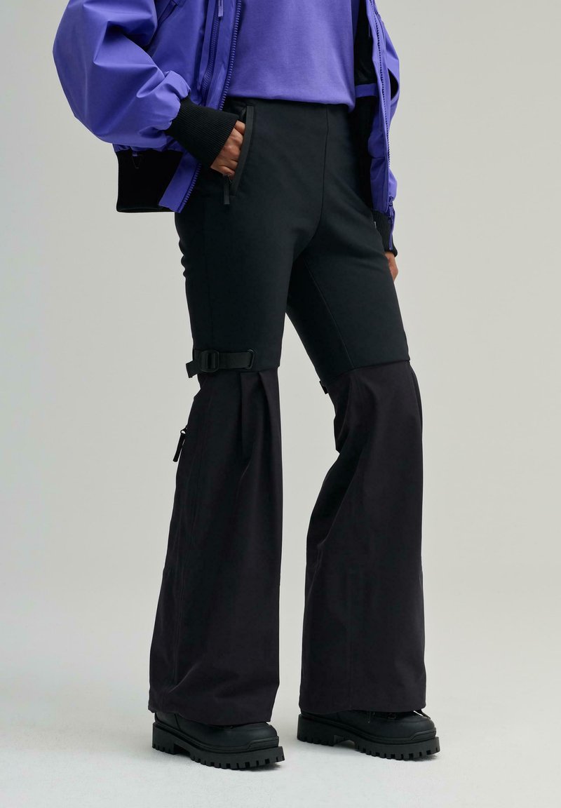 Schwarze Skihose mit ausgestellten Beinen, verstellbaren Trägern an der Taille, Reißverschlusstaschen und glatter Oberfläche, kombiniert mit schwarzen robusten Stiefeln.
