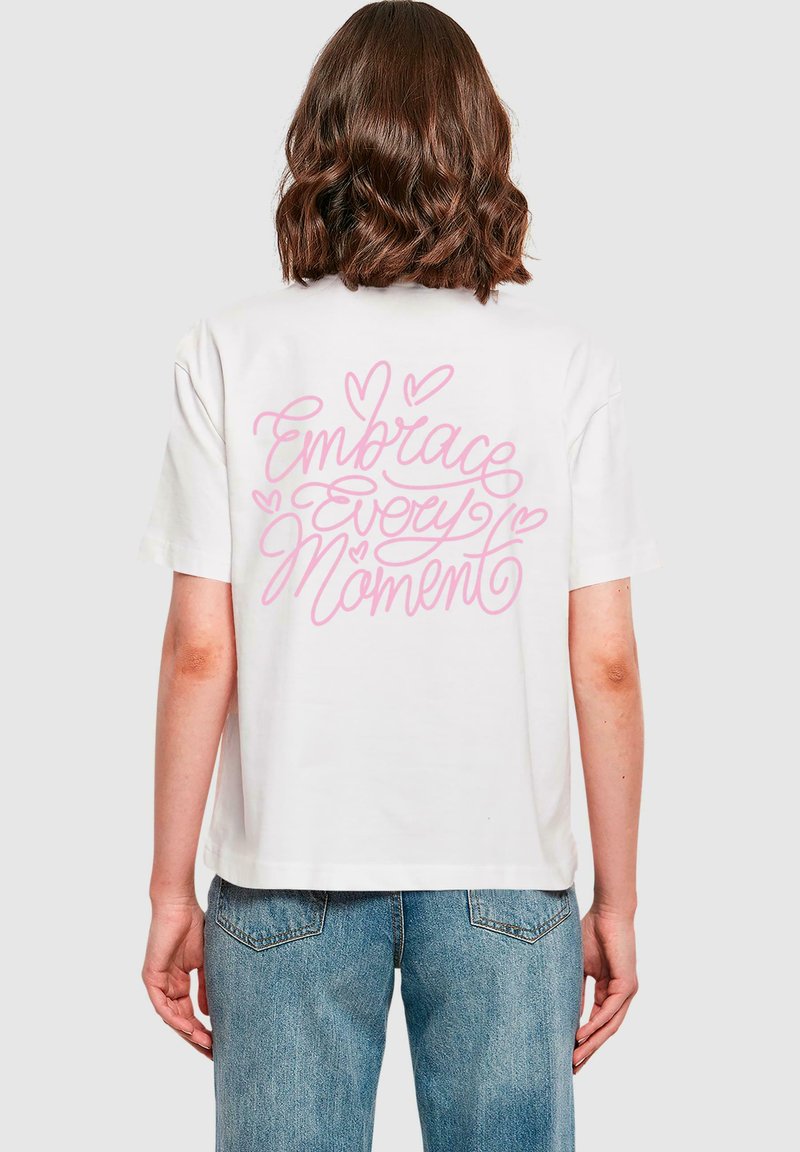 Vit bomulls t-shirt med en avslappnad passform. Bakom finns pink cursiv text "Omfamna Varje Ögonblick" omgiven av hjärtan. Klassisk rund hals.
