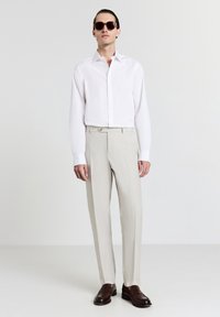 Chemise blanche à manches longues, pantalon beige clair avec un bouton, et chaussures brunes foncées ; le modèle porte de grandes lunettes de soleil brunes.