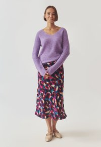 Femme portant un pull en V lavande, une jupe midi florale colorée et des ballerines beiges, se tenant les mains jointes devant un fond uni.