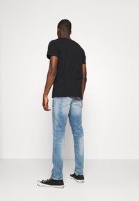 Camiseta negra de algodón, jeans ajustados azul, zapatillas negras. La modelo está de espaldas sobre un fondo blanco liso, destacando los detalles del atuendo.