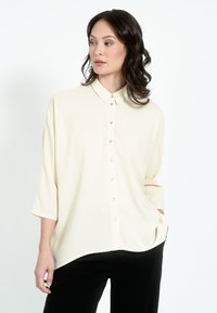 Kreemika värvi pluus kraega, esiosal nööbid ja kolmeveerand varrukad. Valmistatud siledast kangast, lõdva lõikega, kombineeritud mustade pükstega.