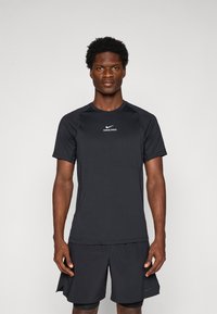 Mann som står vendt fremover iført en svart Nike Pro kortermet skjorte og svarte idrettsshorts mot en enkel bakgrunn.