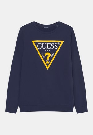 Granatowy długi sweter z żółtym trójkątnym logo z napisem "GUESS U.S.A.", znakiem zapytania oraz słowami "washed jeans."