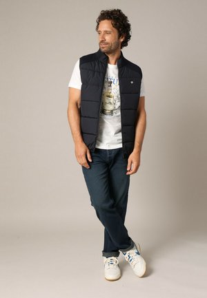 Gilet matelassé bleu marine sur un t-shirt graphique blanc, associé à un jean en denim foncé et des baskets blanches. Design simple avec un tissu léger.